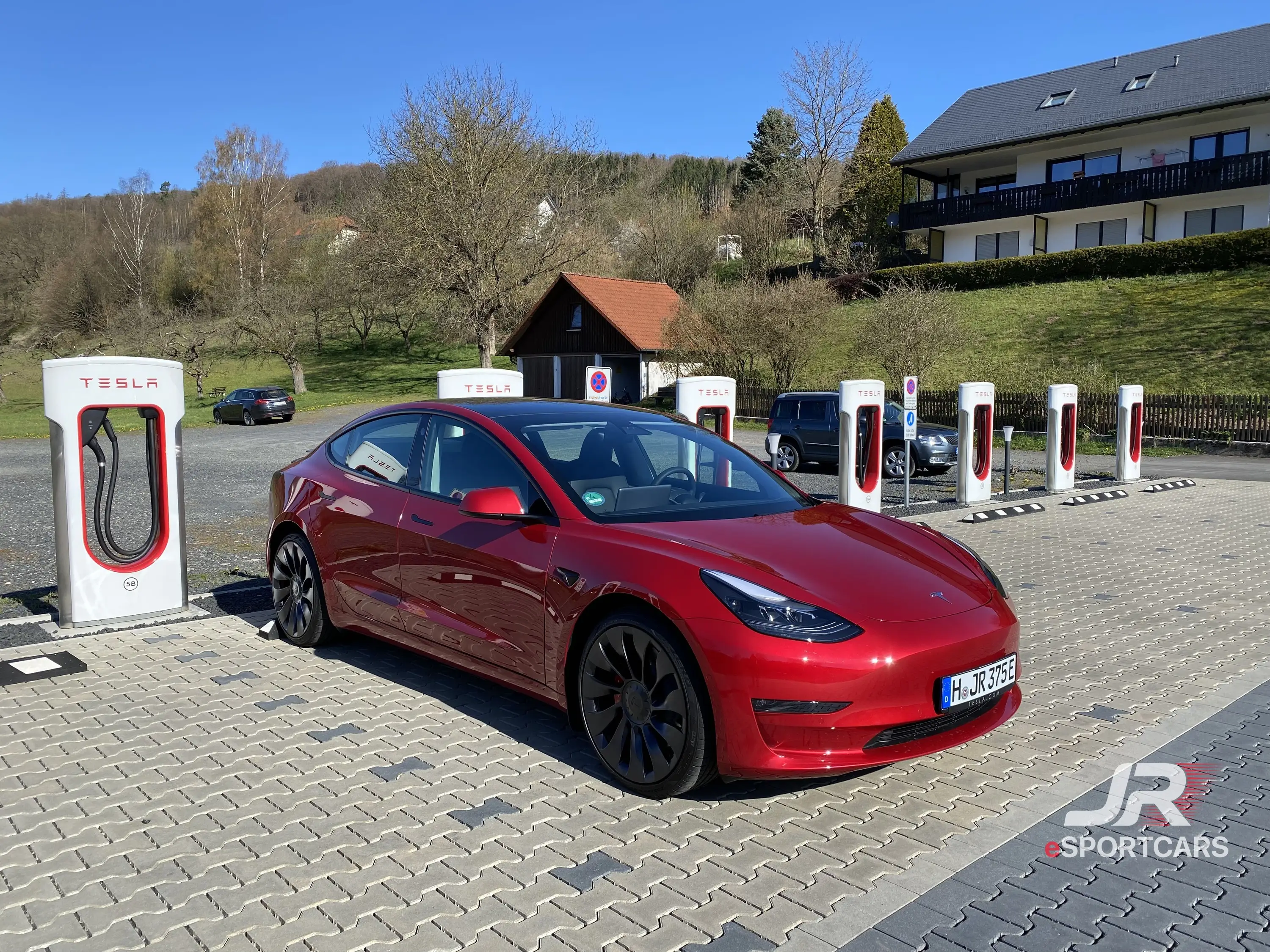 Rotes Tesla Model 3 Performance „Red Devil“, Schrägansicht von vorn, an einem Tesla-Supercharger.