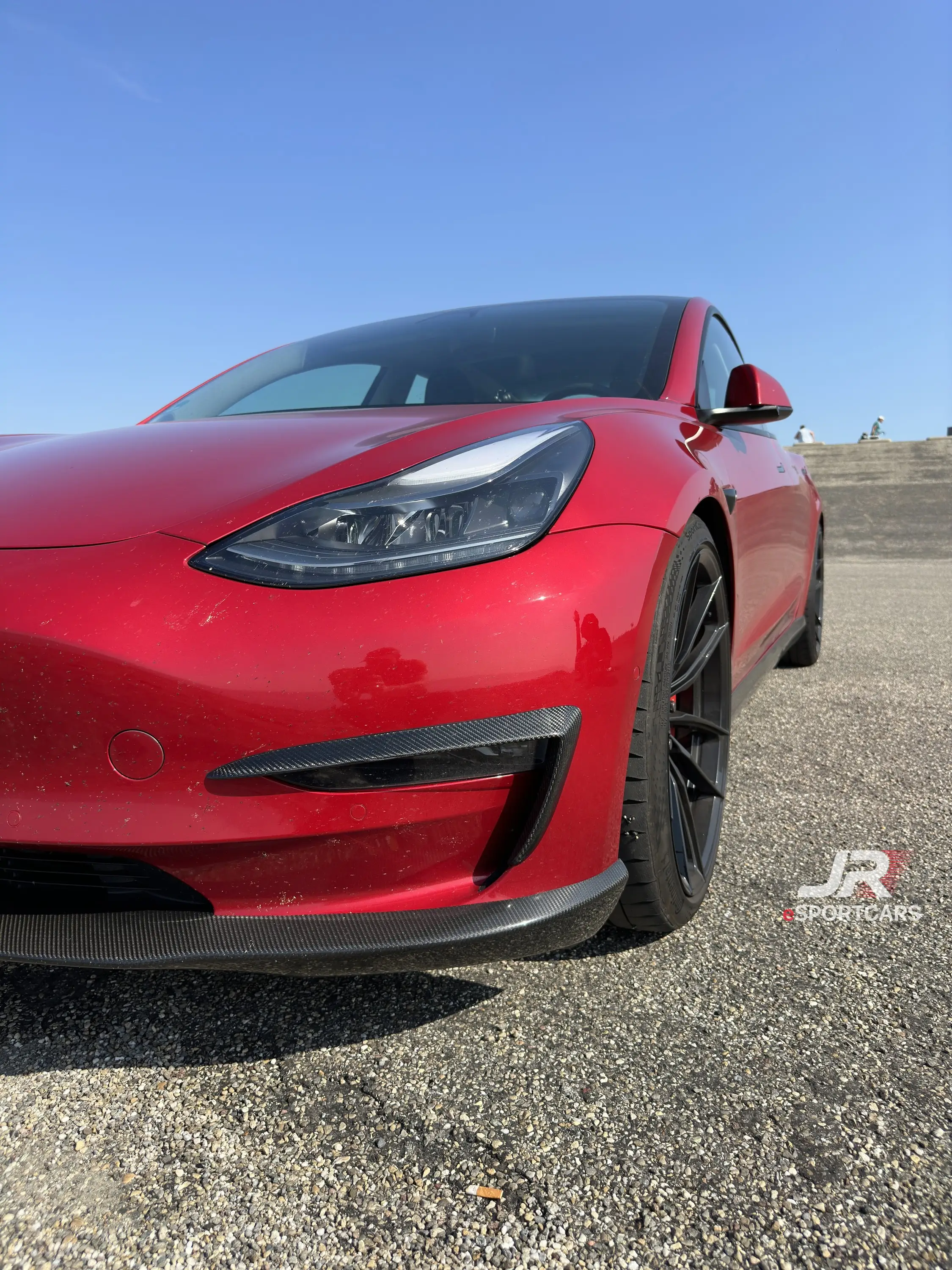 Rotes Tesla Model 3 Performance „Red Devil“, Schrägansicht von vorn mit schwarzer Frontlippe, auf Schotter.