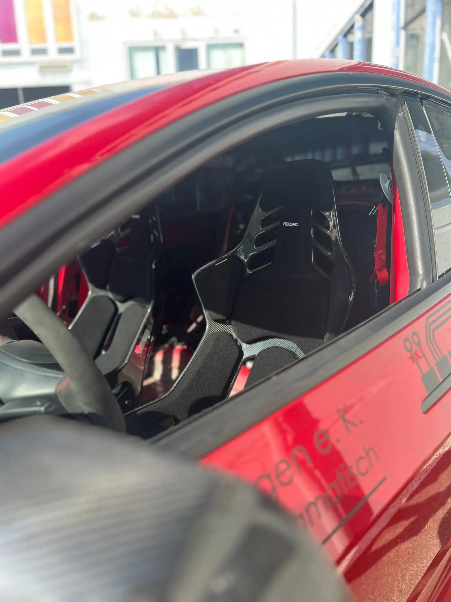 Blick durchs Seitenfenster auf einen schwarzen Recaro-Schalensitz mit roten Gurten.
