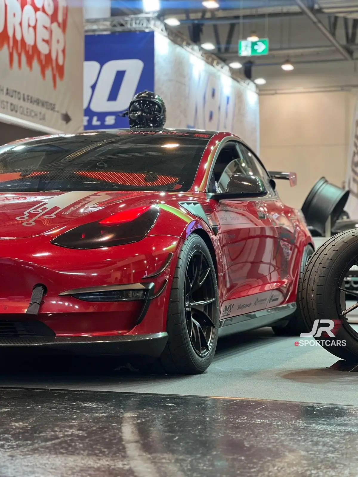 Rotes Tesla Model 3 Performance „Red Devil“, Schrägansicht von vorn in einer Messehalle, ein Helm steht auf dem Dach.