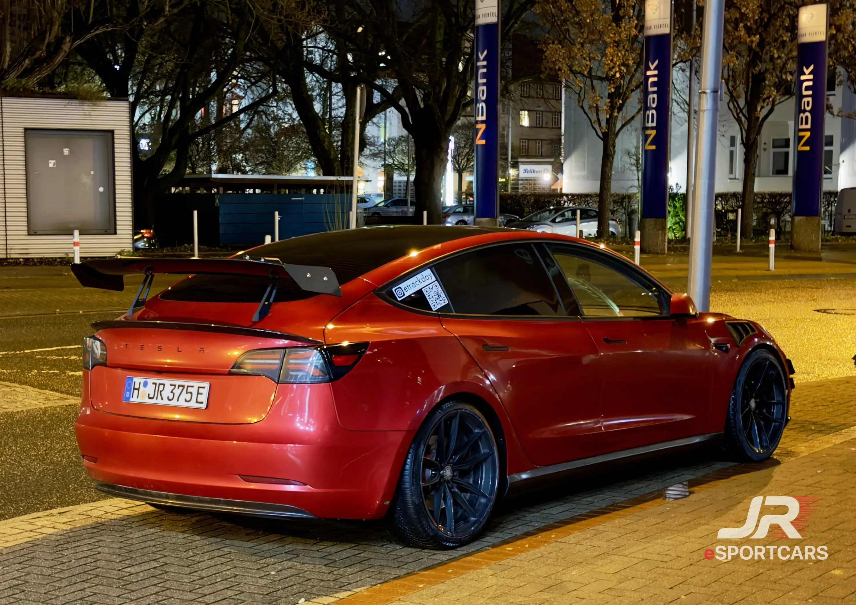 Rotes Tesla Model 3 mit Heckflügel, Schrägansicht von hinten, abends auf einem Parkplatz.
