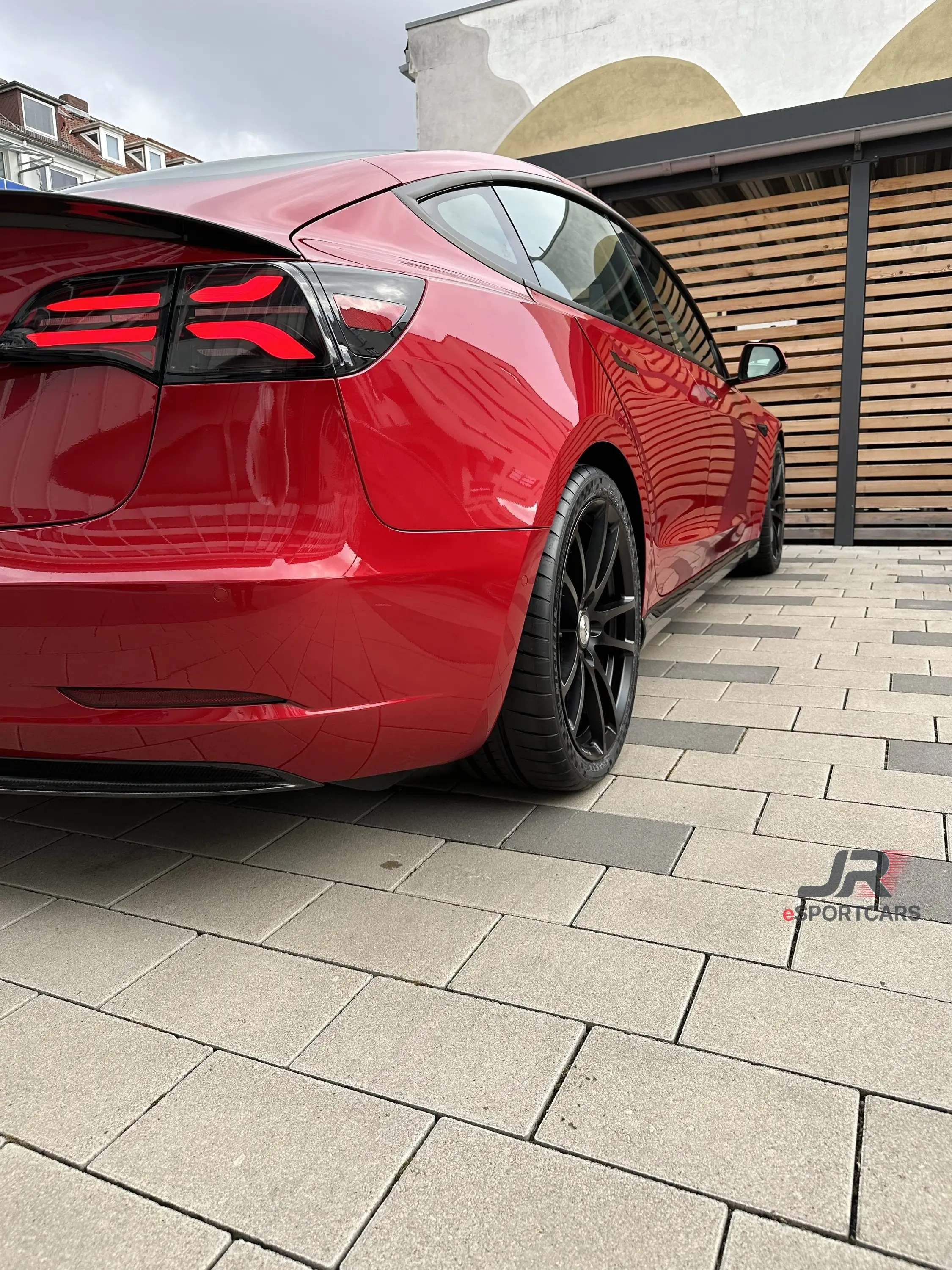 Rotes Tesla Model 3, Schrägansicht von hinten mit dunklen Rückleuchten und schwarzen Felgen.