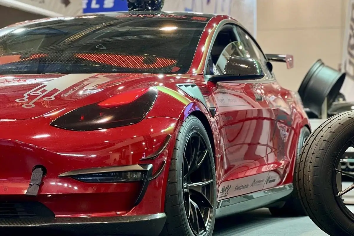 Rotes Tesla Model 3 Performance „Red Devil“ in einer Messehalle, Schrägansicht von vorn, daneben ein Reifen.