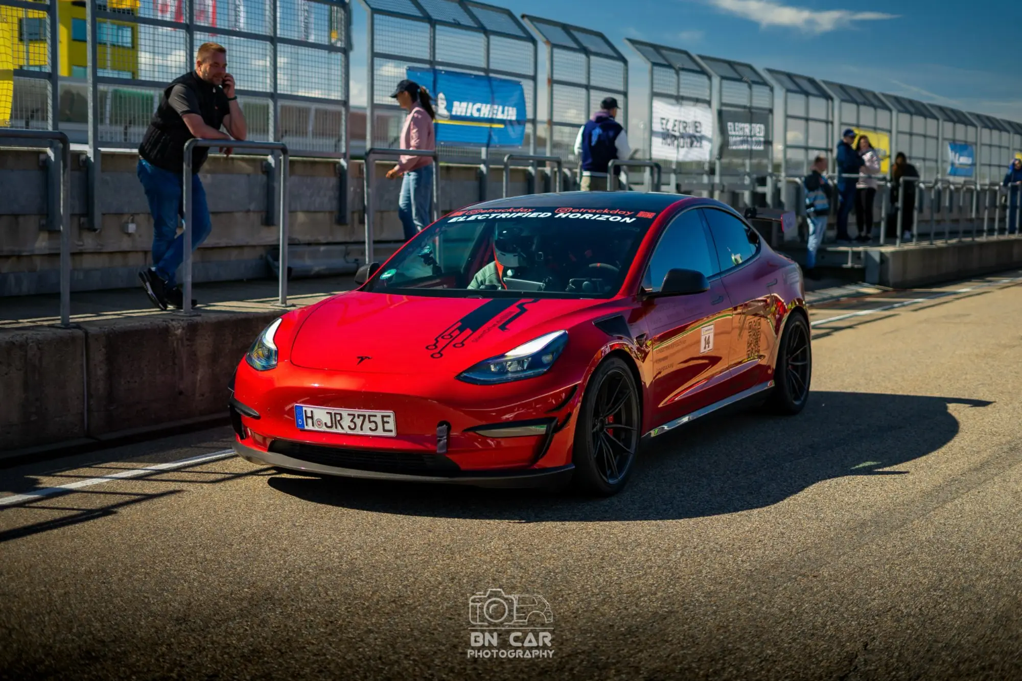 Rotes Tesla Model 3 Performance „Red Devil“ auf der Rennstrecke, Schrägansicht von vorn, Boxenmauer und Zuschauer, Wasserzeichen „BN Car“.